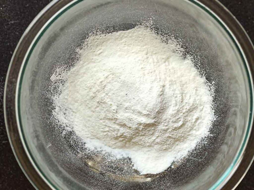 A glass bowl contains a mound of white rice flour powder on a dark surface.